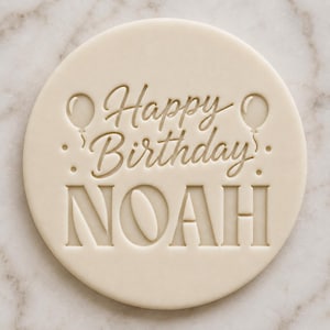 May include: A round, off-white cookie stamp featuring the embossed text Happy Birthday in a cursive script, with the name NOAH in large, bold, sans-serif capital letters below. Two small balloon icons flank the text, with dots.