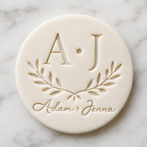 May include: A circular off-white disc featuring an embossed design. The centre displays the letters A and J separated by a dot, positioned above a laurel leaf motif. Below the leaves, the names Adam + Jenna are engraved in a cursive script.