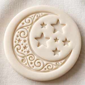 May include: A circular off-white biscuit stamp featuring a debossed design of a crescent moon decorated with intricate swirls and small stars, alongside several larger five-pointed stars positioned within the moon's curve on a flat surface.