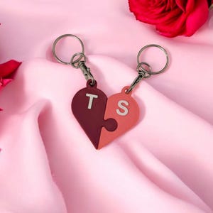 May include: Two silver keychains with a red and pink heart-shaped charm. The heart is split into two puzzle pieces, each with a letter, "T" and "S", on the front.