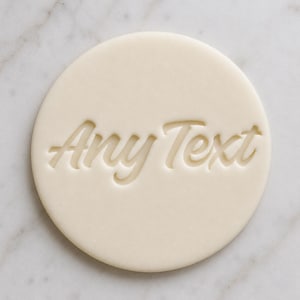 May include: A circular, cream-coloured object with the words "Any Text" embossed in a cursive font. The object is a light beige colour and appears to be made of a smooth material. The background is a light grey and white marble pattern.