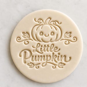 May include: A circular cookie cutter in a pale beige colour, featuring a smiling pumpkin design and the words "Little Pumpkin". The design includes leaves and swirls, ideal for autumn baking.