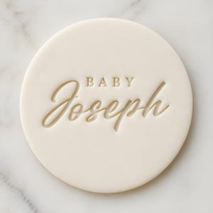 May include: A circular, cream-coloured disc with the word "BABY" above the name "Joseph" in a cursive font. The text is debossed into the surface. The disc is set against a white marble background.