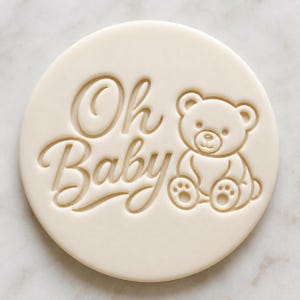 May include: A circular off-white biscuit stamp featuring an embossed design of a seated teddy bear next to the text Oh Baby in a cursive script font. The stamp has a smooth, flat surface with clean, recessed lines forming the bear and lettering.