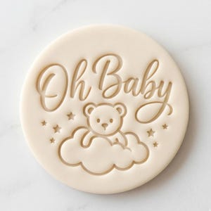 May include: A circular off-white biscuit stamp featuring the embossed text Oh Baby in a cursive script. Below the text is a centred illustration of a teddy bear sitting on a cloud, surrounded by several small five-pointed stars.