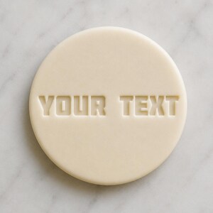 May include: A circular, pale beige cookie with the words "YOUR TEXT" embossed in a blocky font. The cookie is set against a white and grey marble surface.