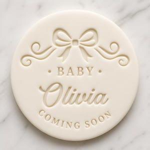 May include: A circular, cream-coloured disc featuring embossed text and a bow motif. The text reads "BABY Olivia COMING SOON". A bow is at the top, with decorative swirls. The disc is set against a marble background.