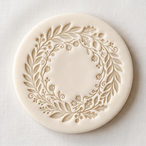 May include: A circular off-white silicone mould featuring a recessed botanical wreath design. The pattern consists of intricate leaves, small berries, and delicate flower buds arranged in a continuous ring around a smooth, flat centre.