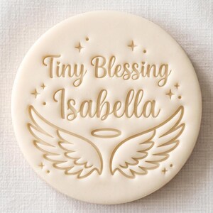 May include: A round, off-white cookie stamp featuring the embossed text "Tiny Blessing Isabella" in a cursive font. Below the text, there is a graphic of two spread angel wings and a small halo centered above them, surrounded by tiny star accents.