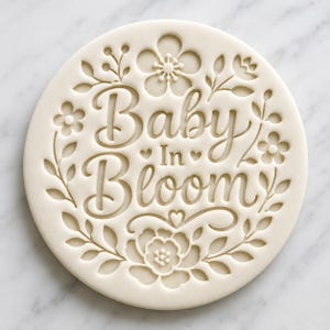 May include: A round, off-white cookie cutter with the words "Baby In Bloom" in elegant script. The text is surrounded by floral and leaf designs, including flowers and hearts. The cutter is made of a light-colored material.