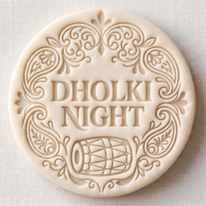May include: A circular off-white embossed biscuit or stamp featuring the text DHOLKI NIGHT in the centre. The design includes an intricate paisley border and a small dholak drum icon positioned below the text, all rendered in a relief style.