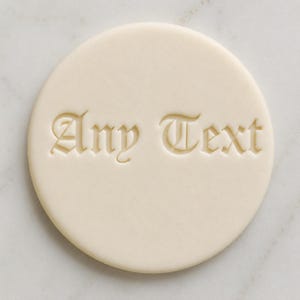 May include: A circular, cream-coloured object with the words "Any Text" embossed in a gothic font. The text is centered on the circular surface. The object is set against a light-coloured background.