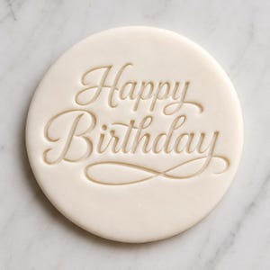 May include: A circular, off-white biscuit stamp featuring the embossed text Happy Birthday in a cursive script font. The design includes a decorative flourish underneath the word Birthday, creating a clean, pressed impression on the flat surface.