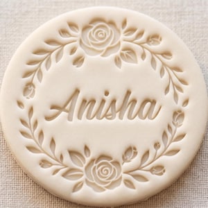 Peut inclure: Un tampon à biscuit circulaire blanc cassé présentant le nom Anisha en relief au centre. Le texte est entouré d'une couronne décorative composée de roses détaillées, de feuilles et de petits boutons de fleurs gravés sur la surface.