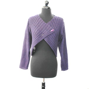 May include: A purple knitted cropped sweater with a criss-cross front and a small pink button detail.