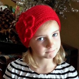 May include: A young girl wearing a red knitted headband with a large red flower accent. The headband is crocheted and has a simple design. The girl is wearing a black and white striped shirt.