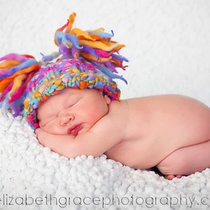 May include: A newborn baby wearing a colorful, handmade knit hat with vibrant orange, yellow, blue, and purple accents. The baby is lying on a soft, white textured blanket, peacefully asleep. The image is a professional photograph.