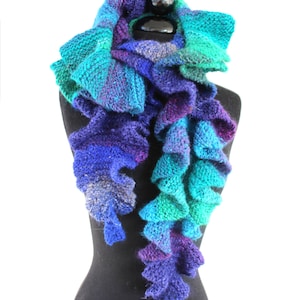 Puede incluir: Una bufanda de ganchillo en tonos de turquesa, morado y azul con una textura rizada. La bufanda se muestra en un maniquí negro.