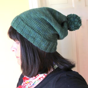 Puede incluir: Un gorro de punto verde oscuro con un pompón a juego. El gorro es holgado y tiene un borde de canalé.