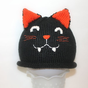 May include: A black knitted hat in the shape of a cat's head, with orange ears, white embroidered eyes and teeth, and an orange nose. The hat has a brim and is designed to be worn on the head. The hat is likely for a child.