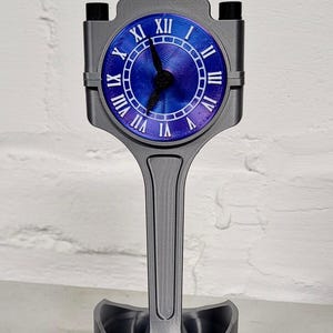 May include: A silver-colored clock with Roman numerals on a blue face, mounted on a silver-colored engine piston. The piston is attached to a silver-colored base with a black ring around the bottom.