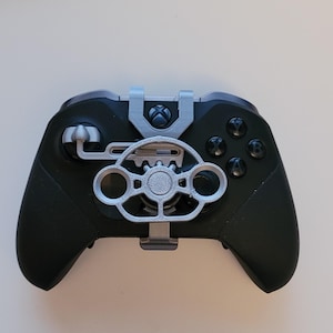 May include: A black Xbox controller with a silver 3D printed steering wheel attachment. The steering wheel has a gear design and two side buttons.