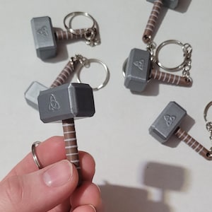 May include: Miniature gray hammer keychains with brown handles and silver key rings. Each hammer features a Celtic knot design on the head. Several keychains are scattered on a white surface.