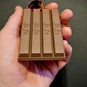 May include: A brown chocolate bar ornament with the text "Kit Kat" printed on each of the four sections. The ornament is attached to a red and green ribbon.