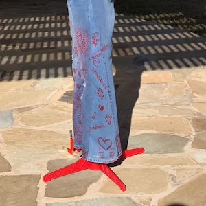 May include: Blue denim jeans with red painted designs, including hearts, flowers, and swirls. The jeans are displayed on a red tripod stand.