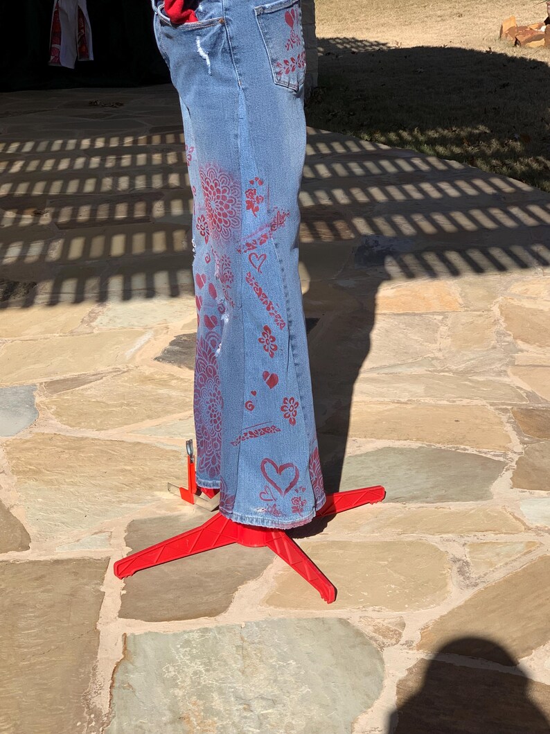 May include: Blue denim jeans with red painted designs, including hearts, flowers, and swirls. The jeans are displayed on a red tripod stand.