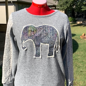Könnte beinhalten: Ein graues Sweatshirt mit einem großen Elefanten-Applikationen auf der Vorderseite. Der Elefant ist aus einem gemusterten Stoff gefertigt und hat einen weißen Spitzenbesatz. Die Ärmel des Sweatshirts sind ebenfalls mit weißer Spitze besetzt.