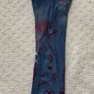 May include: A pair of blue denim jeans with a distressed look and red floral designs. The jeans have a wide leg and a low rise.