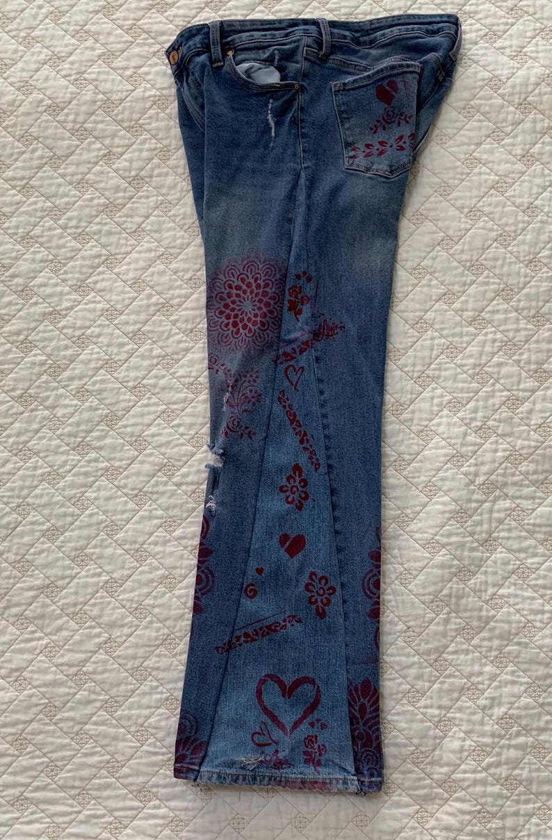 May include: A pair of blue denim jeans with a distressed look and red floral designs. The jeans have a wide leg and a low rise.