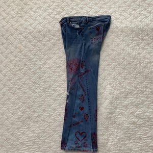 May include: A pair of blue denim jeans with a red and white pattern. The jeans have a flared leg and are decorated with hearts, flowers, and other designs.
