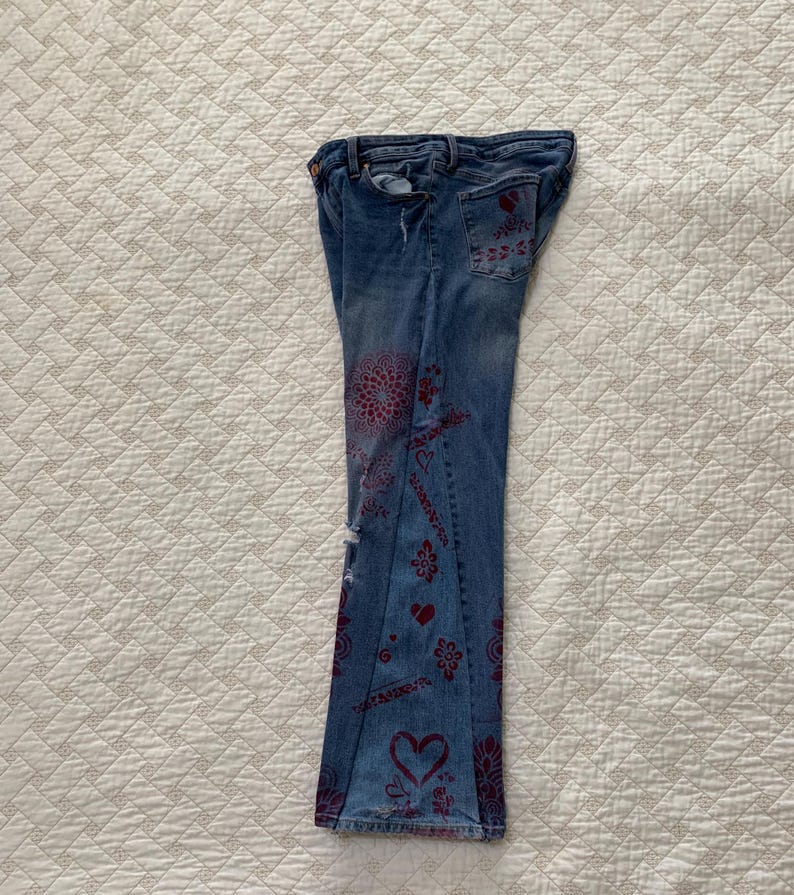 May include: A pair of blue denim jeans with a red and white pattern. The jeans have a flared leg and are decorated with hearts, flowers, and other designs.