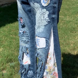 May include: Distressed denim jeans with floral patterns and patches. The jeans have a white fabric panel sewn down the side seam.