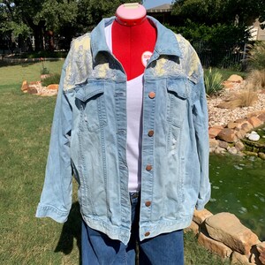 May include: A light blue denim jacket with a white and blue floral pattern on the shoulders. The jacket has distressed details and a button closure.