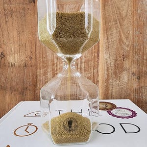 May include: A glass hourglass with gold sand, sitting on a white surface with the text "The Good Lab" printed on it. The hourglass is shaped like a diamond and has a clear glass body.
