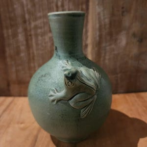 May include: A green ceramic vase with a frog sculpted on the side. The vase has a round body and a narrow neck.