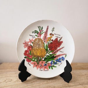 Puede incluir: Un plato decorativo blanco con un diseño floral colorido, ilustraciones botánicas y texto. El plato se exhibe sobre un soporte negro. Las flores incluyen flores rojas, rosas, amarillas y azules.