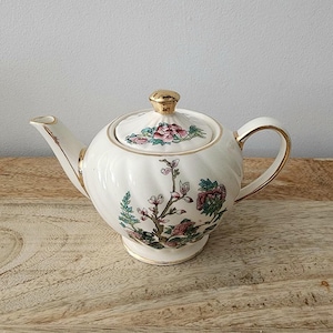 Può includere: Teiera in ceramica color crema con un design floreale. La teiera presenta un manico, un beccuccio e dettagli del coperchio color oro. Il coperchio è sormontato da un pomello dorato. Il design floreale include fiori e foglie rosa, rosse e verdi.