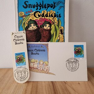 May include: A vintage book titled "Snugglepot and Cuddlepie" with a blue cover and illustrations of two children. The book is accompanied by two stamps featuring the same characters, one with a 33c denomination.