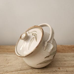 May include: A round, off-white ceramic sugar bowl with a matching lid and a single arched handle. The surface features hand-painted green leaf motifs and brown rim detailing. The bowl has a smooth, glazed finish and a sturdy, rounded base.