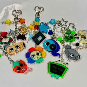 Puede incluir: Una colección de llaveros de acrílico con varios personajes y objetos de dibujos animados, incluyendo una flor, un televisor, un casete y una fresa. Cada dije está unido a un broche plateado en forma de corazón con cuentas de colores.