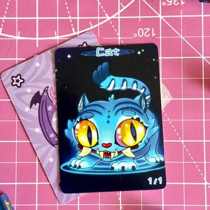 May include: A fantasy-themed trading card featuring a blue cat with large yellow eyes and sharp teeth. The card is labeled "Cat" and "1/1" on a dark blue background with stars. A second card with a purple bat illustration is also visible.