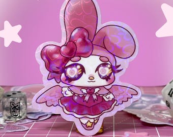 May include: A holographic sticker featuring a cartoon character with bunny ears, heart-shaped eyes, and wings. The character is dressed in a pink dress with a bow. The sticker has a shiny, iridescent finish and is set against a pink background with star accents.