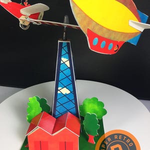 May include: A vintage paper model featuring a red biplane with a pilot, a yellow and red airship, a blue skyscraper, a red house, and green trees. The model is set on a green base with red star-shaped flowers. The Paper Retro logo is visible.