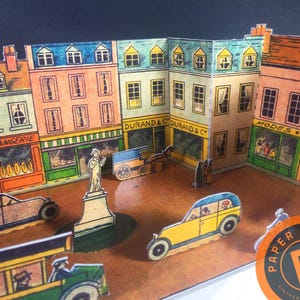 May include: A paper diorama depicting a European street scene with colorful buildings, shops, and vintage vehicles. The buildings feature signs such as "Boulangerie" and "Durand & C". Includes a statue and the "Paper Retro" logo.