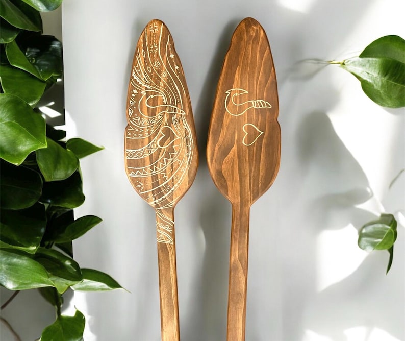Op de afbeelding: Twee houten peddels met ingewikkelde ontwerpen erin gegraveerd. De peddels zijn bruin en hebben een lichtbruin ontwerp. De ontwerpen zijn van een vogel en een hart.
