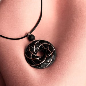 May include: A black pendant necklace with a circular design featuring a rose gold wire pattern. The pendant is hanging on a black cord.